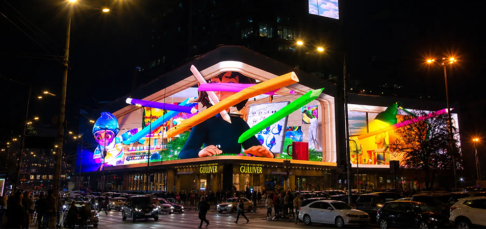 3D billboard2 LED Display Manufacturer in China HonghaiLED Nighttime city scene with a large building projection featuring oversized colorful glowing pencils and pedestrians nearby near a Gulliver store façade