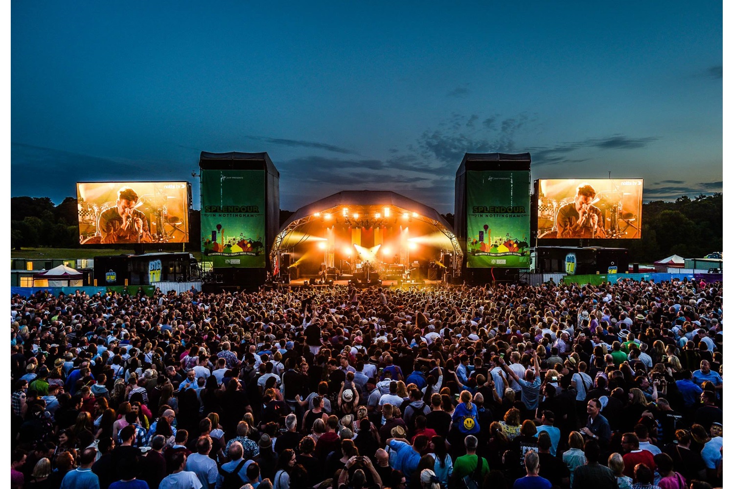 Outdoor music festival crowd facing a bright central stage with two large video screens at duskearly nightfall